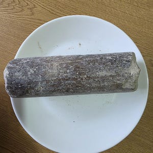 Puede incluir: Un objeto cilíndrico de color marrón oscuro con una textura rugosa, posiblemente de madera, descansa sobre un plato blanco. El plato está centrado en una superficie de madera. Los extremos del objeto son irregulares y la apariencia general sugiere un material natural.