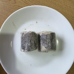 Może przedstawiać: Dwa ciemnoszare, prostokątne obiekty o szorstkiej fakturze leżą na białym talerzu. Obiekty wydają się być produktami spożywczymi, prawdopodobnie brykietami węgla drzewnego lub podobną substancją. Talerz stoi na jasnobrązowej drewnianej powierzchni.