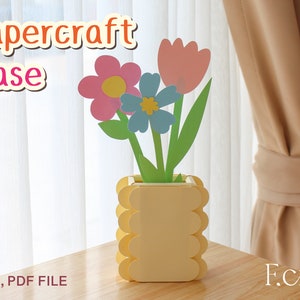 Puede incluir: Jarrón de papel con el texto "Papercraft Vase" en la parte superior de la imagen. El jarrón es amarillo y tiene un diseño festoneado. Está lleno de tres flores de papel: una flor rosa, una flor azul y un tulipán rosa.