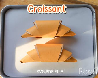 May include: Two paper croissants, with a light brown color, are arranged on a metal tray. The word "Croissant" is written in orange above the pastries. Below, the text "SVG, PDF FILE" is visible.