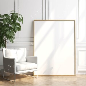 May include: A contemporary interior scene with a large, blank white framed canvas. A white armchair with a square cushion and a green plant in a pot are in the foreground. The floor is made of light-coloured wood. The wall is white.