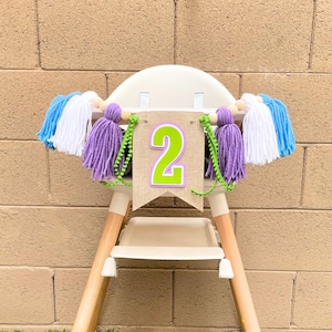 May include: A white high chair with a burlap banner that says "2" in green letters. The banner is decorated with purple and white pom-poms and blue and white streamers.