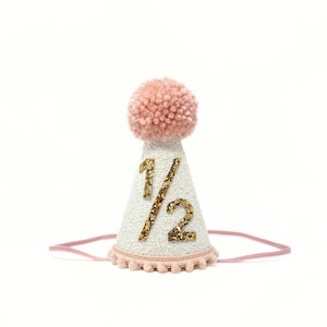 May include: A white party hat with a pink pom-pom on top. The hat has gold glitter numbers that say "1/2" on the front.