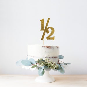 May include: A white cake with gold glitter frosting and a gold glitter cake topper that says "1/2". The cake is on a white cake stand and is surrounded by green eucalyptus leaves.