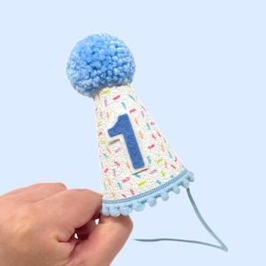 May include: A blue and white birthday hat with a blue pom-pom on top and a number one on the front. The hat has a white background with colorful sprinkles and a blue trim.