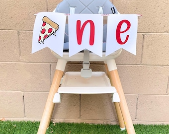 May include: A high chair with a banner that says "one." The banner has white pennants with red letters and a pizza slice graphic. The high chair has a gray seat, white tray, and wooden legs.