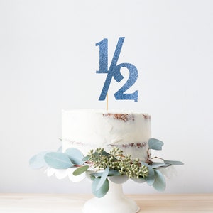 May include: A white cake with blue glitter frosting and a blue glitter cake topper that says "1/2". The cake is on a white cake stand and is decorated with green eucalyptus leaves.