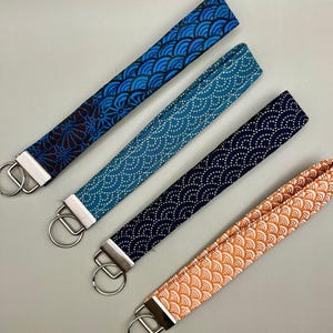 May include: Four fabric key fobs in shades of blue and orange, each with a different patterned design. The key fobs have a silver metal clasp and ring. The patterns include a wave design and a dotted design.