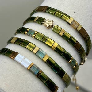 May include: Several beaded bracelets in shades of green, gold, and white are displayed. One bracelet features a gold star charm. The bracelets are made of small, rectangular beads and are strung together with a delicate cord.