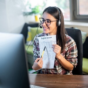 May include: A woman wearing a headset and glasses is teaching an English lesson online. She is holding a sheet of paper with the words "English lesson" written on it and pointing to a section with a pen.
