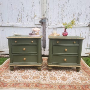 May include: Two olive green nightstands with three drawers each, featuring gold-toned round knobs and decorative gold trim along the edges and base. Each nightstand stands on four short, rounded feet and has a smooth, matte finish.