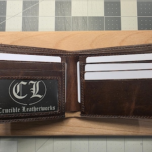 May include: A brown leather bifold wallet featuring multiple card slots and a clear ID window. The ID window contains a black insert with the white text CL and Crucible Leatherworks. The wallet is open, displaying its interior compartments.