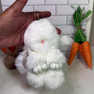 May include: A fluffy white bunny plush toy with a pink nose and gold accents on its paws. The bunny is held in a hand. Decorative orange carrots with green tops are in the background.