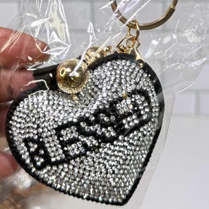 May include: Heart-shaped keychain with the word "BLESSED" spelled out in black letters. The heart is covered in sparkling rhinestones and has a gold-colored key ring and charm.