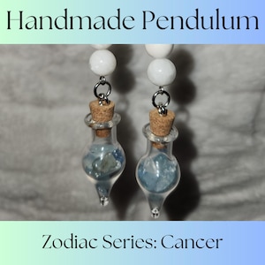 May include: Handmade pendulum earrings with a white bead and a glass bottle filled with blue crystals. The bottle has a cork stopper and is attached to a silver chain. The earrings are labeled "Zodiac Series: Cancer".