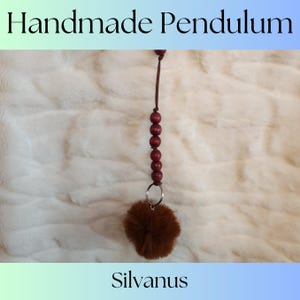 May include: Handmade pendulum with a brown fur pom-pom and a string of maroon wooden beads. The pendulum is hanging from a brown leather cord. The background is a white faux fur.
