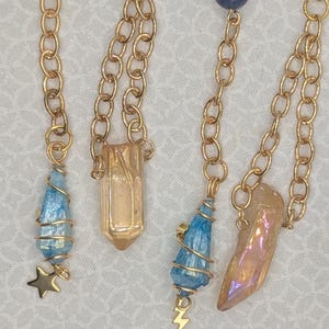 May include: Four gold chains with different charms. The charms are a blue teardrop, a clear teardrop, a gold teardrop, and a clear teardrop. The chains are all connected to a gold ring.
