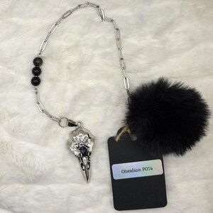 May include: A silver-toned chain necklace featuring three dark obsidian beads, a silver bird skull pendant engraved with a compass design, and a large black faux fur pom-pom. A black rectangular tag is attached, displaying the text Obsidian P014.