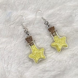 May include: A pair of dangle earrings featuring clear, star-shaped glass bottles filled with bright yellow glitter. Each bottle is sealed with a small brown cork stopper and attached to a silver-toned metal fishhook ear wire for hanging.