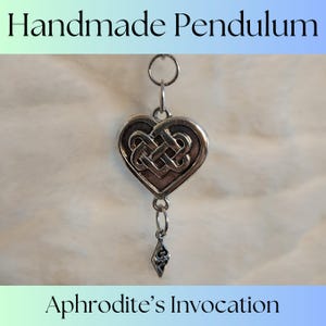 May include: A silver heart-shaped pendulum with a Celtic knot design and a smaller silver charm hanging below. The pendulum is labeled "Aphrodite's Invocation".