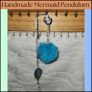 May include: Handmade mermaid pendulum with a blue pom-pom, white beads, a silver chain, and a silver charm with a mermaid design.