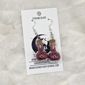May include: Sterling silver dangle earrings featuring miniature glass bottles filled with dark pink glitter and sealed with cork stoppers. The earrings are displayed on a white card with a black cat and moon graphic, plus the text MOONVIXENWITCHERY.