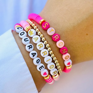 May include: Four bracelets with pink, white and gold beads. The bracelets spell out the words "STRONG", "WARRIOR", "COURAGE", and a heart.