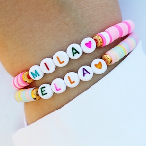 May include: Two bracelets with colorful beads spelling out the names "MILLA" and "ELLA" with a heart charm on each bracelet.