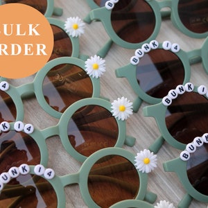 May include: Bulk order of sage green children's sunglasses. Each pair features round lenses, black lettering spelling out 'BUNK 14', and a white daisy flower accent. The sunglasses are made of plastic and appear lightweight and durable, suitable for kids.