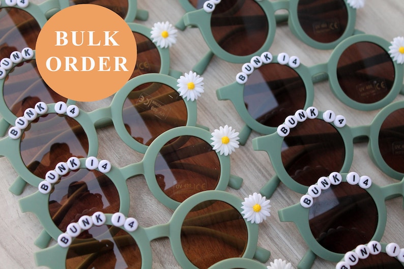 May include: Bulk order of sage green children's sunglasses. Each pair features round lenses, black lettering spelling out 'BUNK 14', and a white daisy flower accent. The sunglasses are made of plastic and appear lightweight and durable, suitable for kids.