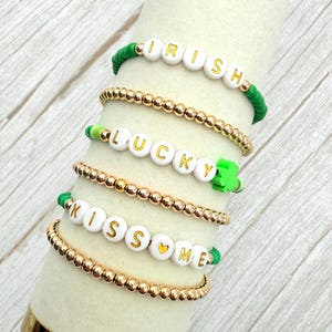 Peut inclure: Cinq bracelets avec des perles dorées et des perles de lettres blanches qui épellent "Irish", "Lucky" et "Kiss Me". Les bracelets sont sur une surface en bois blanc.