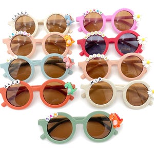 May include: Ten pairs of children's sunglasses with various names and dinosaur accents.  The frames are in assorted colors including pink, red, orange, light green, and beige. Each pair features a child's name spelled out on the front and a small dinosaur charm.  The lenses are brown. These personalized sunglasses are perfect for kids.