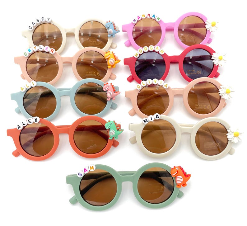 May include: Ten pairs of children's sunglasses with various names and dinosaur accents.  The frames are in assorted colors including pink, red, orange, light green, and beige. Each pair features a child's name spelled out on the front and a small dinosaur charm.  The lenses are brown. These personalized sunglasses are perfect for kids.