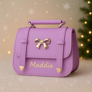 May include: A lavender purse with a gold bow and handle. The purse has a gold clasp and the name "Maddie" in gold lettering. The purse has a structured design with a top handle and shoulder straps.