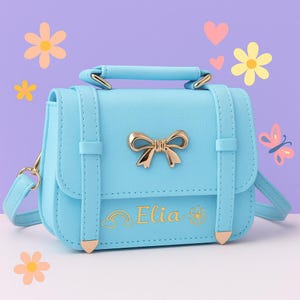 May include: A light blue satchel-style purse with a gold bow clasp and handle. The purse has a gold bow accent and the name "Elia" in gold script with a rainbow and flower graphic. The purse has a shoulder strap.