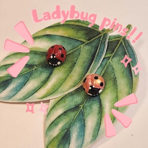May include: Two red and black ladybug pins on a green leaf background. The text "Ladybug pins!!" is written in pink letters above the ladybugs.