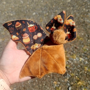May include: A plush bat toy with brown fur and patterned wings. The wings feature a dark background with images of cupcakes, coffee mugs, and autumn leaves. The bat's body is a solid brown color.