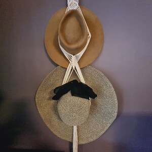 May include: A macrame hat holder displaying two hats. The top hat is brown, and the bottom hat is straw-coloured with a black ribbon. The macrame is cream-coloured and hangs from a white hook.