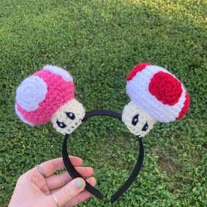 May include: A black headband featuring two crocheted mushroom designs. One mushroom is pink with white spots, and the other is red with white spots. The mushrooms have cream-colored stems and black eyes.