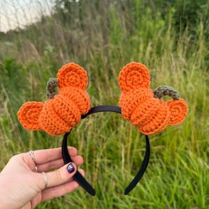 May include: A black headband featuring two crocheted orange pumpkins, each with a small green stem and two rounded orange ears attached to the sides, creating a festive accessory with a textured yarn finish and a simple dark band base.