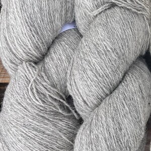 Puede incluir: Primer plano de dos grandes madejas de lana gris. La lana es gruesa y texturizada, con un aspecto suave y natural. La palabra "Grey" está impresa en la parte inferior. Ideal para proyectos de punto y ganchillo.