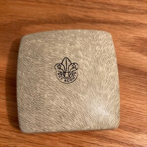 Puede incluir: Una caja blanca cuadrada con una superficie texturizada. La caja tiene un logotipo negro de Boy Scouts of America en la parte superior.