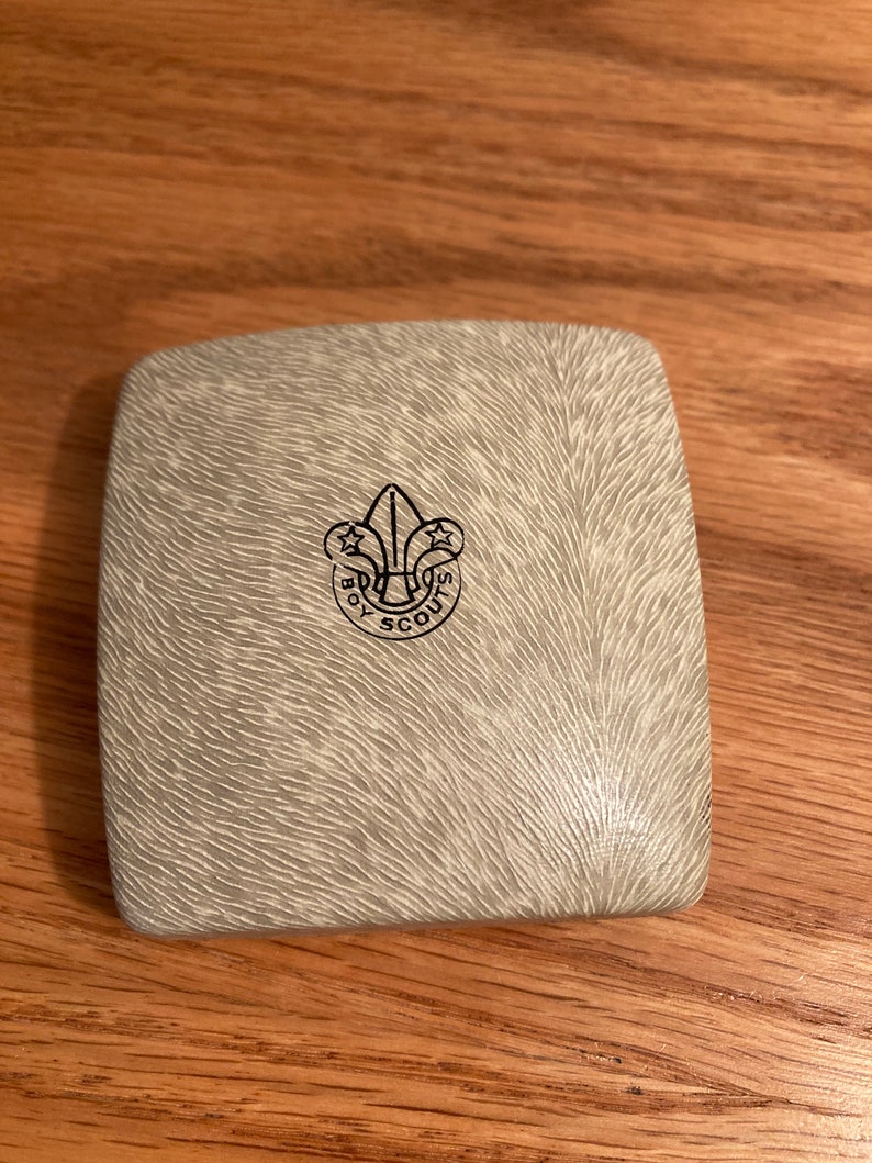 Puede incluir: Una caja blanca cuadrada con una superficie texturizada. La caja tiene un logotipo negro de Boy Scouts of America en la parte superior.