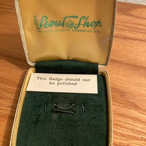 Puede incluir: Una peque&ntilde;a caja rectangular beige con un interior de terciopelo verde. La caja tiene una etiqueta en la parte superior que dice "The Scout Shop, The Boy Scouts Association". La caja tambi&eacute;n tiene una peque&ntilde;a tarjeta en el interior que dice "This Badge should not be polished".