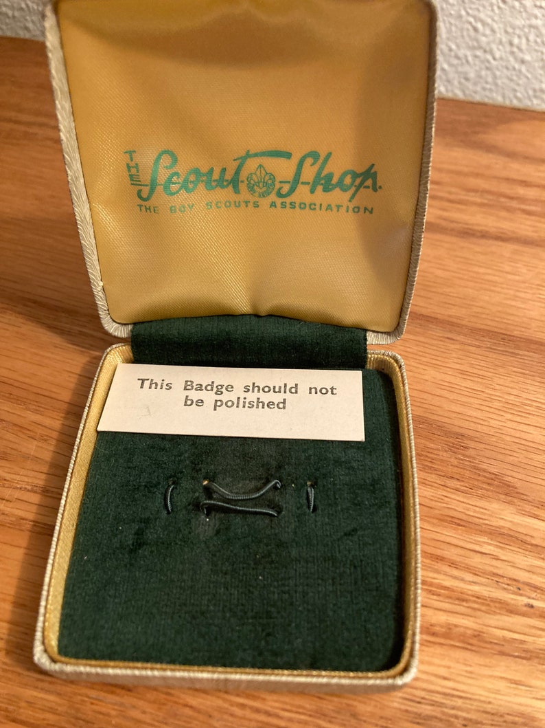 Puede incluir: Una peque&ntilde;a caja rectangular beige con un interior de terciopelo verde. La caja tiene una etiqueta en la parte superior que dice "The Scout Shop, The Boy Scouts Association". La caja tambi&eacute;n tiene una peque&ntilde;a tarjeta en el interior que dice "This Badge should not be polished".