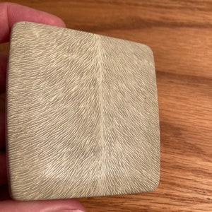 Puede incluir: Una peque&ntilde;a caja cuadrada de color gris claro con una superficie texturizada. La textura se asemeja a un patr&oacute;n de l&iacute;neas que irradian desde el centro.