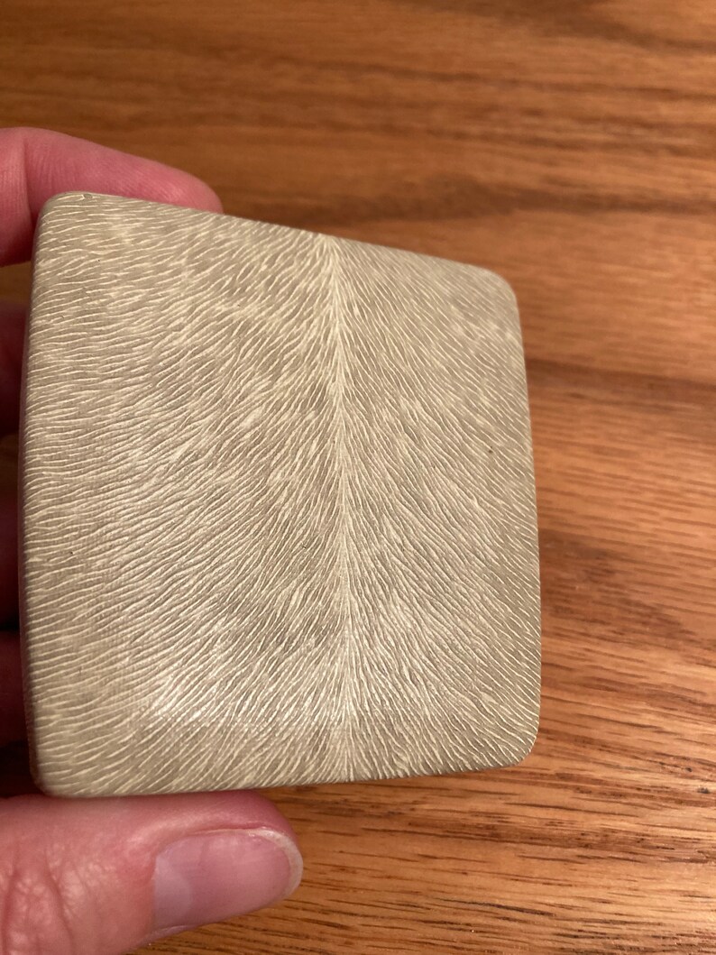 Puede incluir: Una peque&ntilde;a caja cuadrada de color gris claro con una superficie texturizada. La textura se asemeja a un patr&oacute;n de l&iacute;neas que irradian desde el centro.