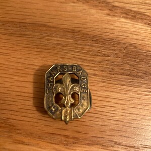 Puede incluir: Un pin de metal dorado con el texto "The Scout Badge" y un dise&ntilde;o de flor de lis.