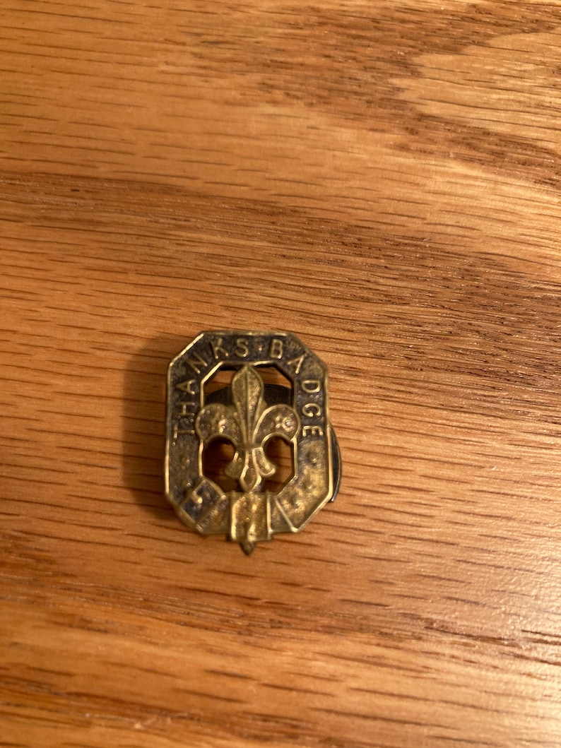 Puede incluir: Un pin de metal dorado con el texto "The Scout Badge" y un dise&ntilde;o de flor de lis.