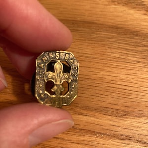 May include: A gold-colored metal pin with a fleur-de-lis design in the center. The pin is surrounded by a circular border with the words 'Thanks for Badge' inscribed around the edge.